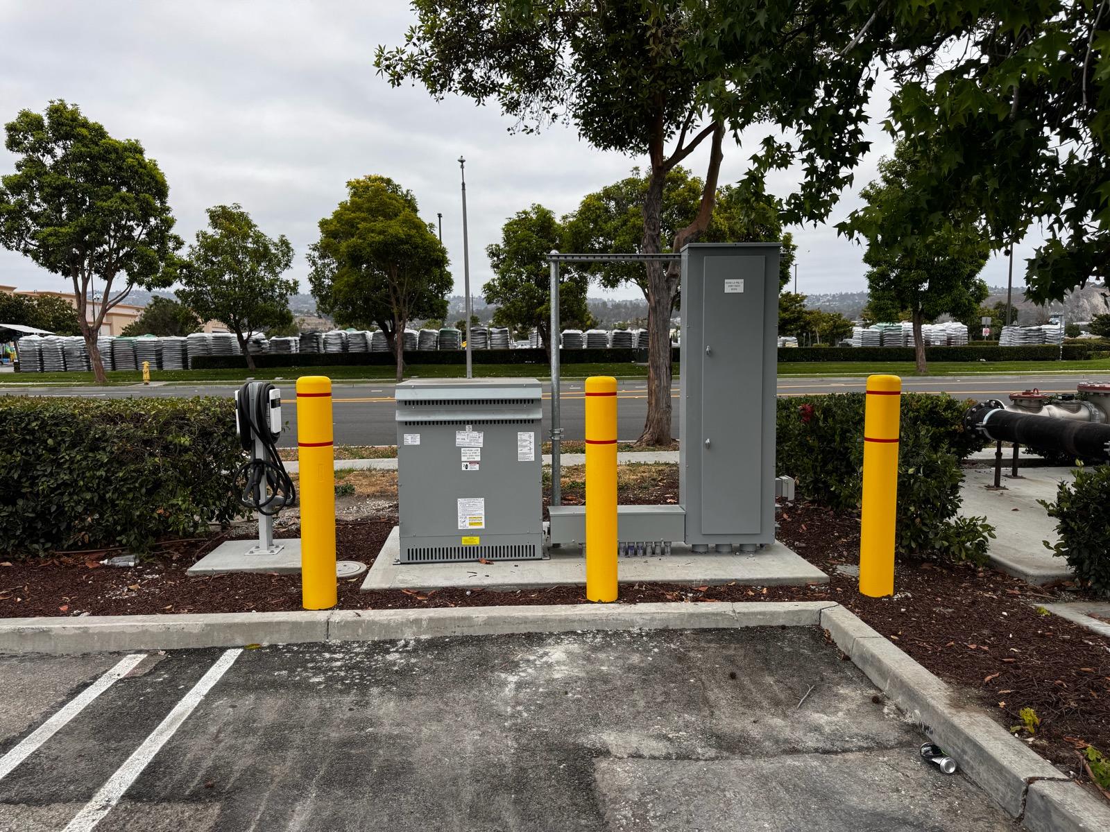 Transformer and panel enclosure with a roadside charging pedestal behind yellow bollards
